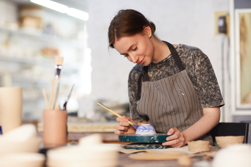 Content attractive skilled young lady in apron standing at table and drawing blue leaves on ceramic bowl in pottery workshop