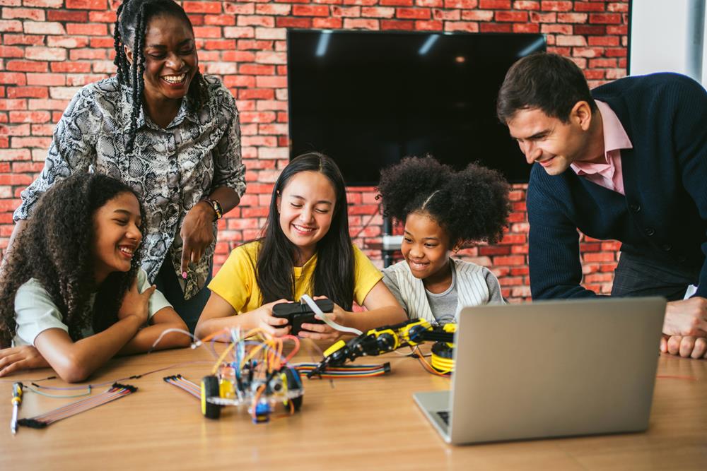 Students and mentors gather around a table with a laptop and electronics kit, smiling as they build and test a small robotics project in a collaborative STEM workshop